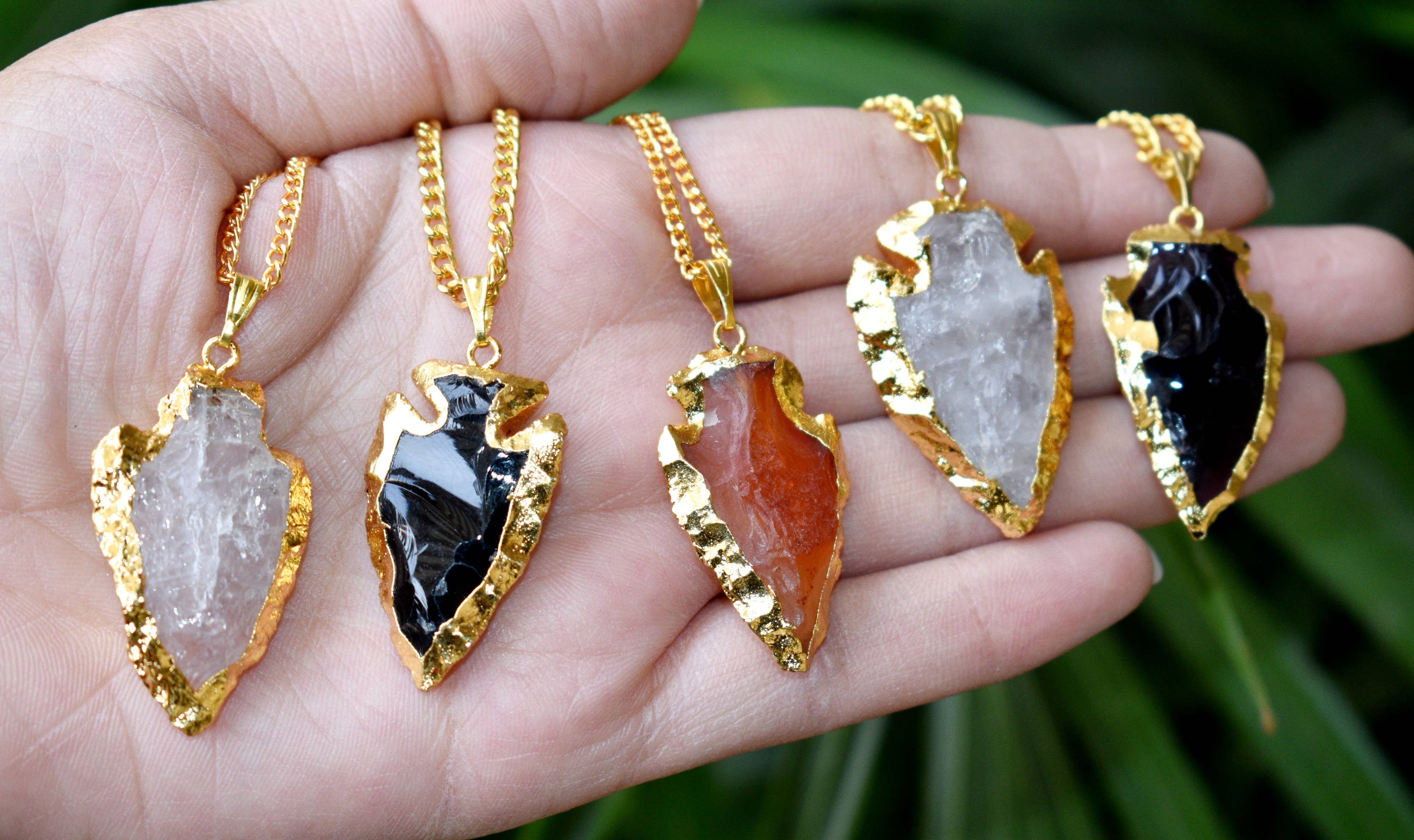 Five arrowhead-shaped pendants on gold chains held in a hand with a natural background.